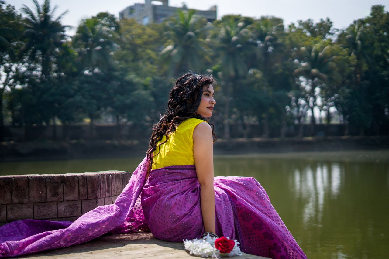 A woman in a purple sari sits by a serene lake, surrounded by lush trees.