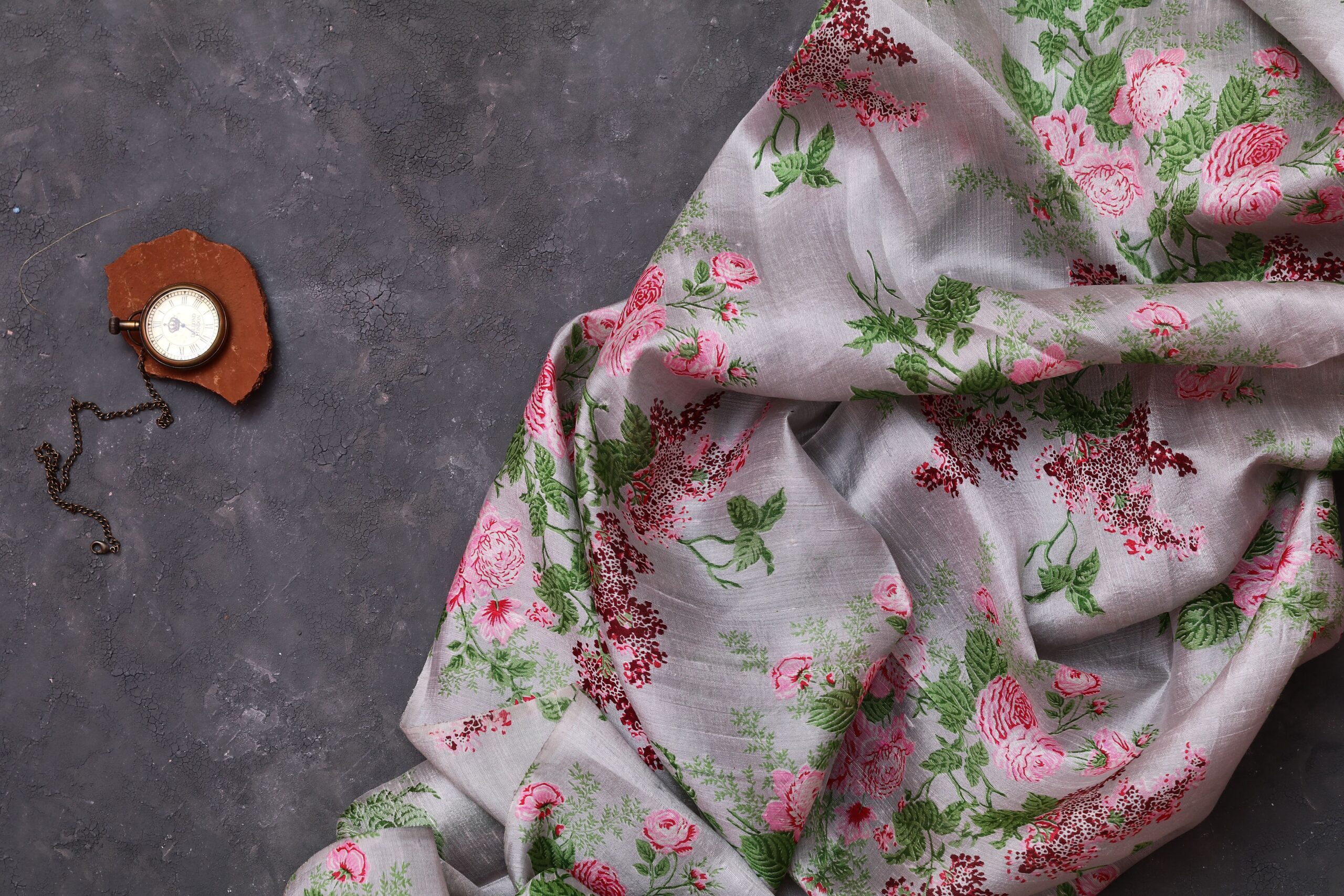 Elegant floral fabric draped beside a vintage pocket watch on a dark surface.
