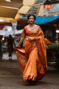 An elegant woman in an orange saree walks confidently through a vibrant outdoor market.
