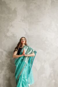Portrait of a graceful Indian woman in a teal saree, hands clasped in a greeting pose.